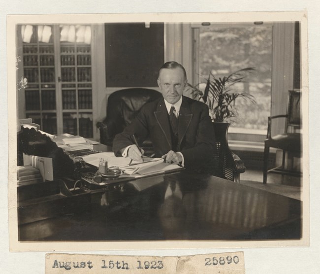 A black and white photo of President Coolidge sitting behind a desk in the Oval Office, holding a pen over paper and looking at the camera.