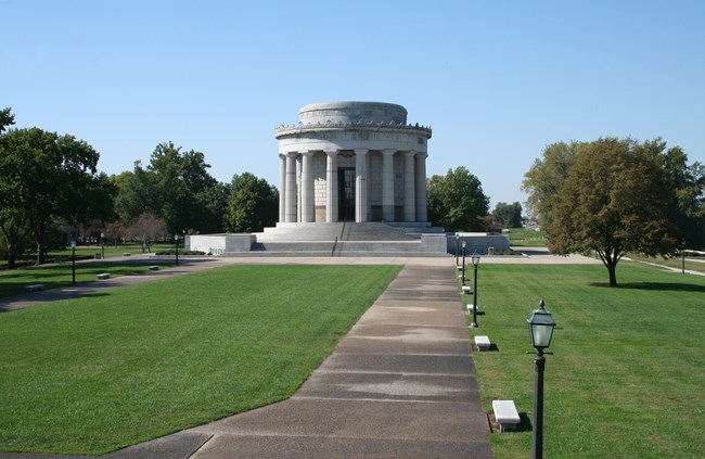 A large gray colonnaded building fronted by stairs. A flat grassy area, sidewalks and small white benches are in the foreground.