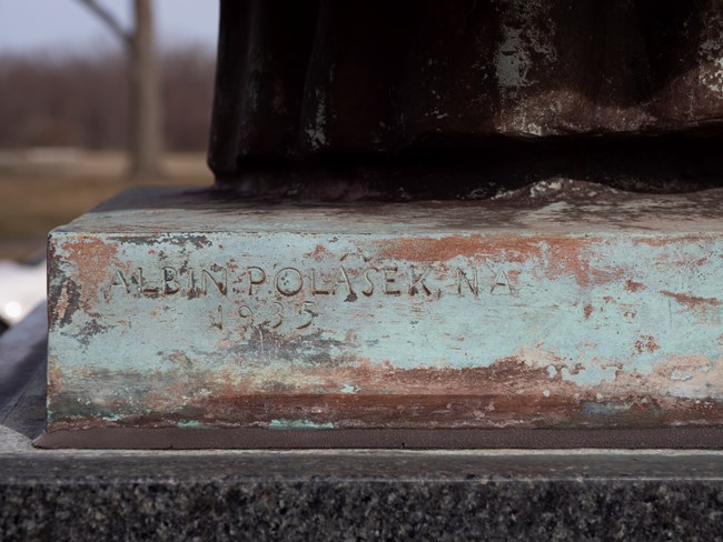 The words Albin Polasek NA stamped in the bronze base of the statue. Albin Polasek is the sculptor NA refers to his membership in the national academy of the arts.