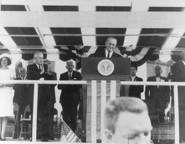 President Lyndon Johnson at a podium with the Seal of the United States on the front. Seven other men stand on the stage along with his wife. Above him is a striped awning, behind them you can see some of the wainscotting on the Clark Memorial building