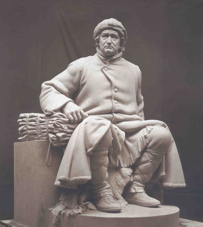 Vigo Statue in studio, a white statue of a man sitting on a bench. He is wearing a long coat, leg gaiters, moccasins and a coon-skin hat. His right arm is bent, elbow on a stack of folded furs and his left arm disappears behind him