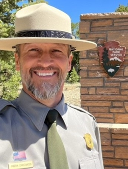 An image of Martin Christiansen a park ranger in grey long sleeved button up shirt, green tie, and felt campaign hat in front of a stone column with the Park Service Arrowhead logo on it.