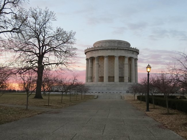 Large, round, gray, colonnaded, building at sunset. the river flowing behind the building reflects the pink and orange hues of the sky. A tree without leaves stands to the left and in front of the memorial.