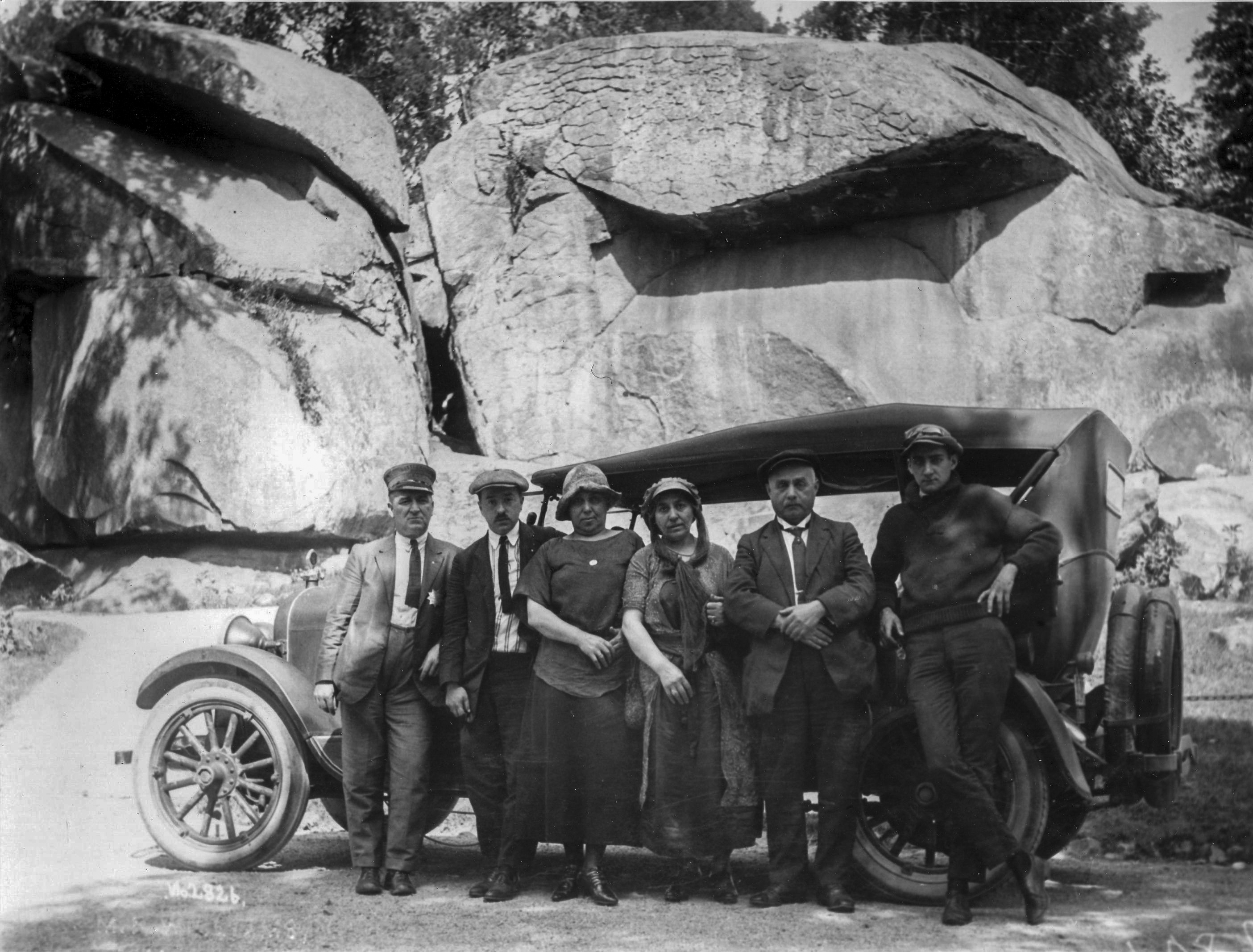 A black and white photo of a group of six adults standing in front of an 1920s era automobile. Behind the group and automobile is a wall of very large boulders.