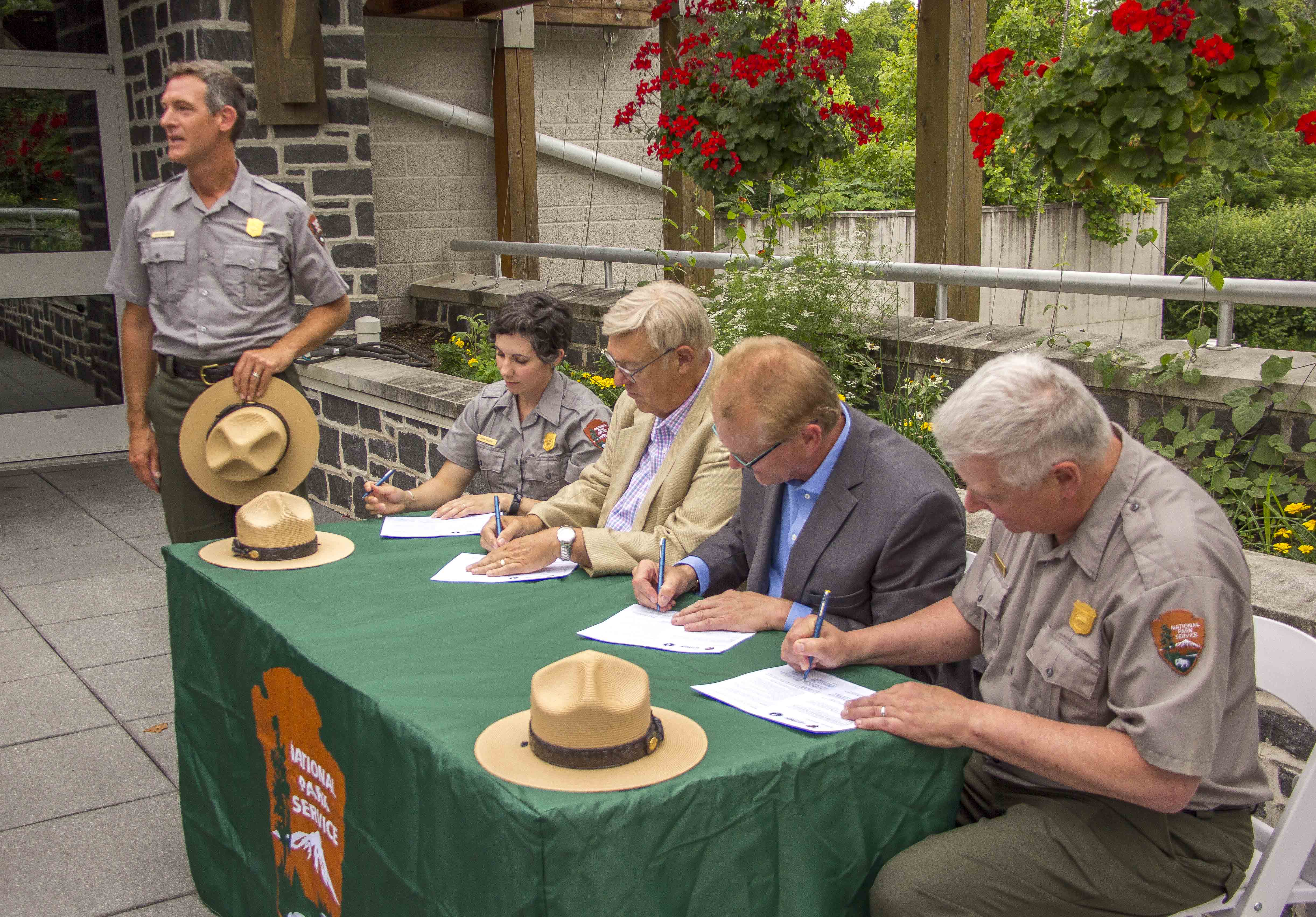 Four people sit at a table as they sign a document while one stands nearby.