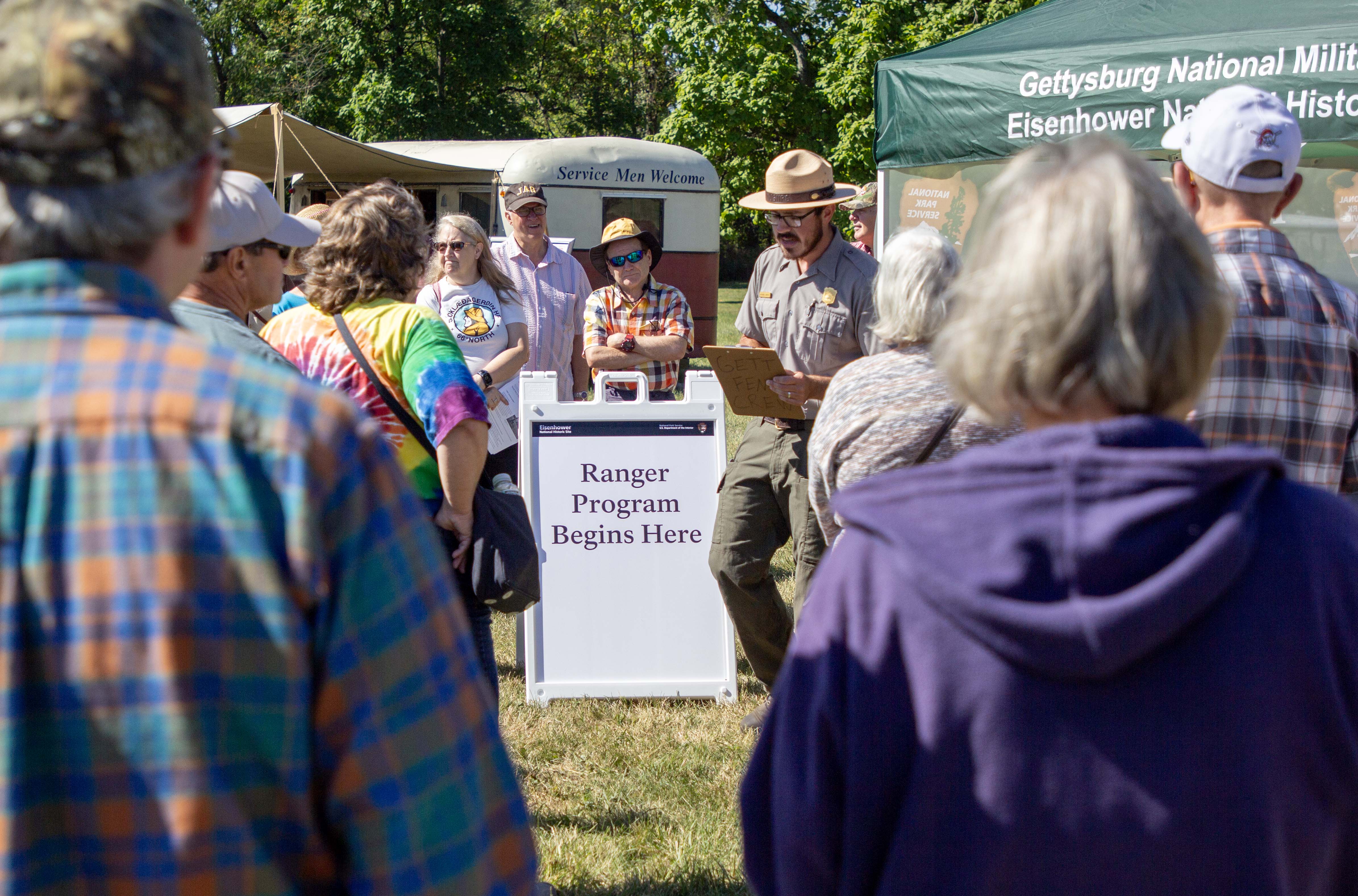 A group of visitors gather around a Park Ranger. In the center is a sign that says Ranger Program Begins Here. Behind the crowd is a red and white trailer that says Service Men Welcome. In the upper right is the top of a green tent.