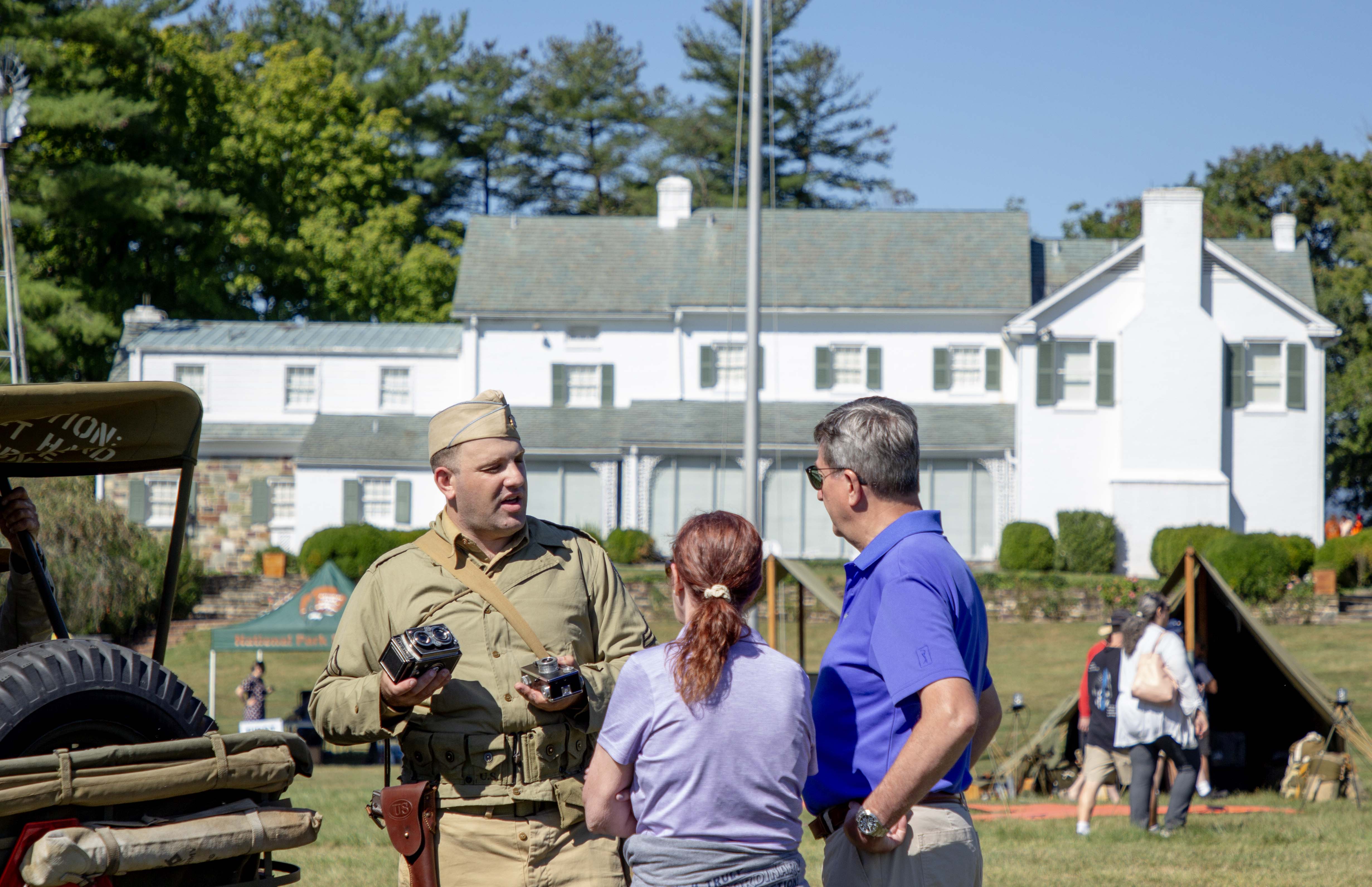 In the foreground is a World War II living historian speaking with two visitors standing next to a Jeep. Behind this group on the right is a khaki colored tent. In the rear is a two-story white house with a green roof and green shutters.