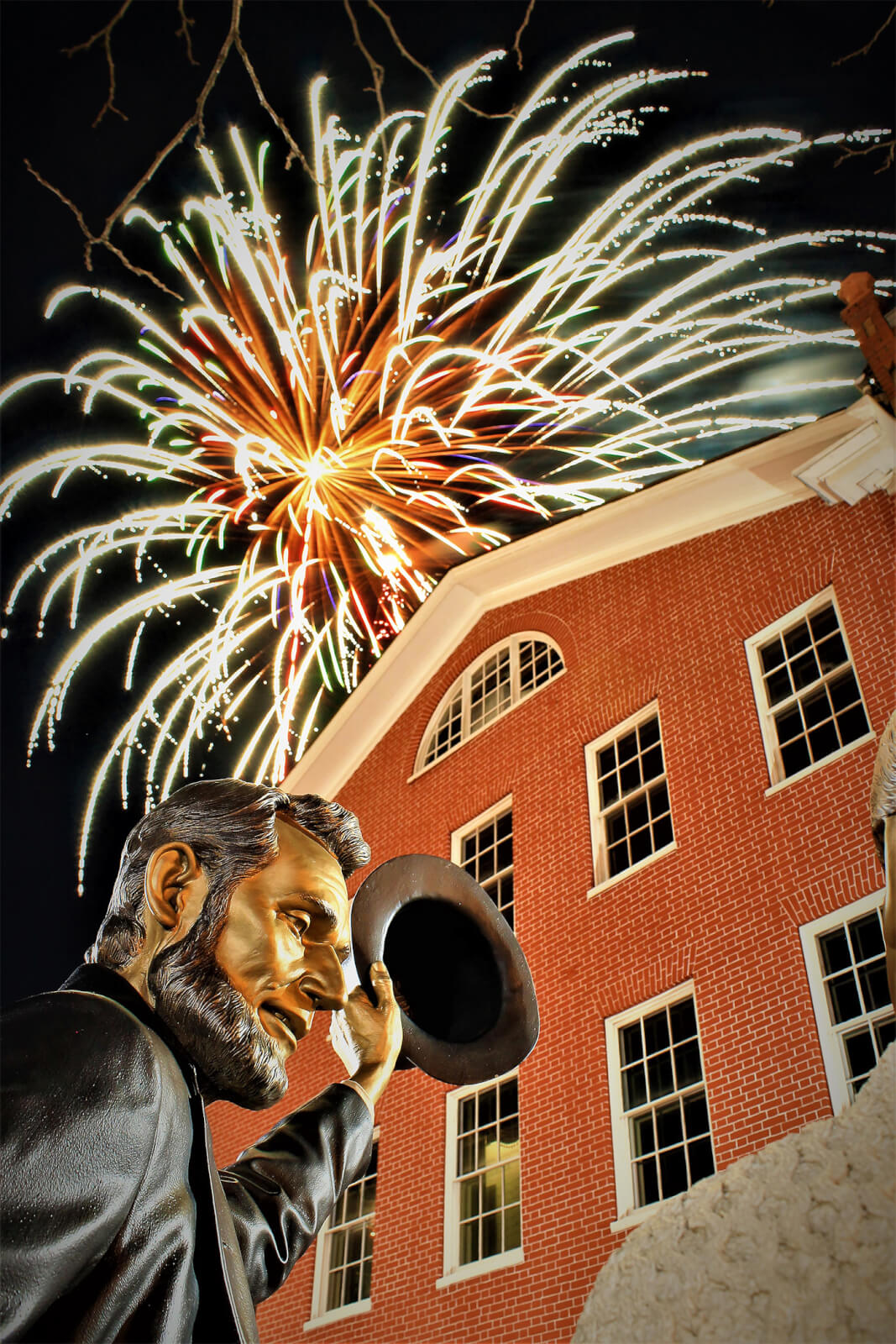 A statue of Abrahm Lincoln stands in front of a three-story red brick house with cream color shutters. In the sky above the building are bright fireworks.