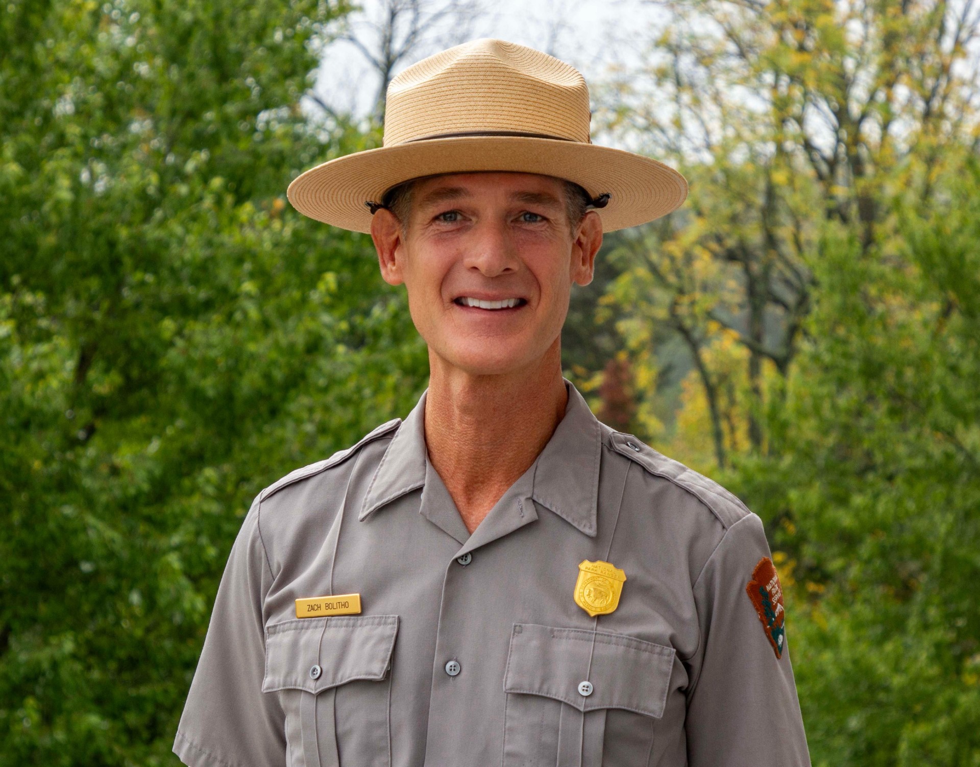 A white male stands for a portrait. He is wearing a grey button-down uniform shirt with a patch on the left sleeve, a gold badge over his left chest, and a gold name bar over his right chest. He is wearing a flat trooper hat.