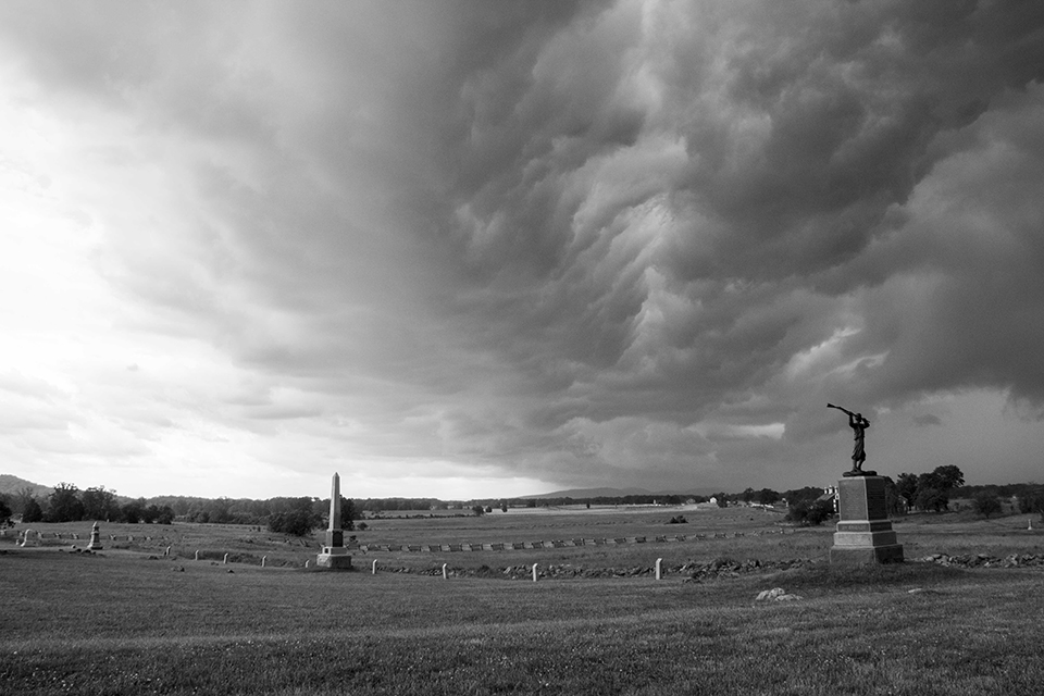 A black and white photo of an open field with two monuments in the foreground and stone and wooden fences in the distance. Storm clouds roll over the area from above.