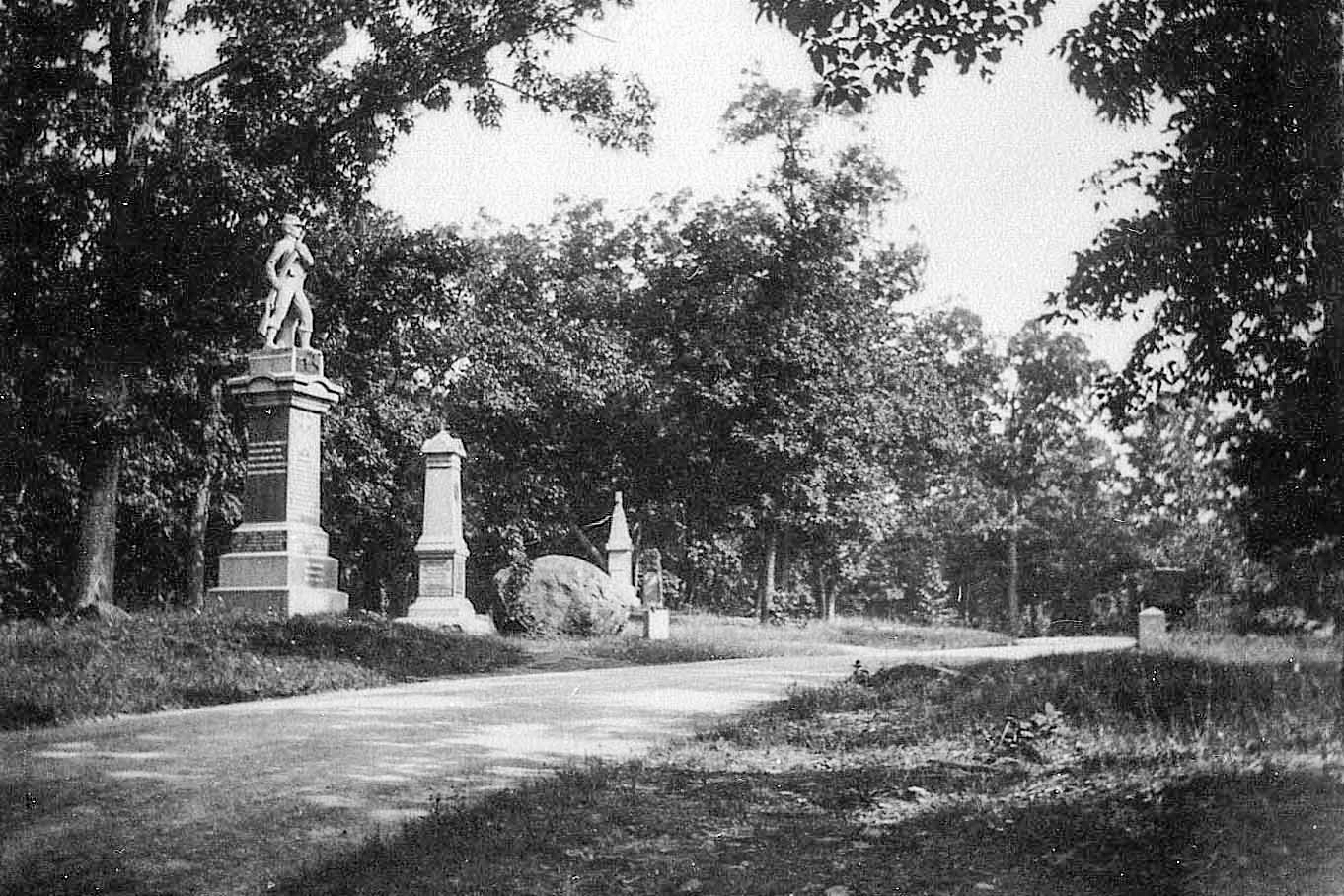 A black and white photo of a wooded scene with a gravel road in the center running left to right. Along the left side of the road is a series of three monuments and a large round boulder.