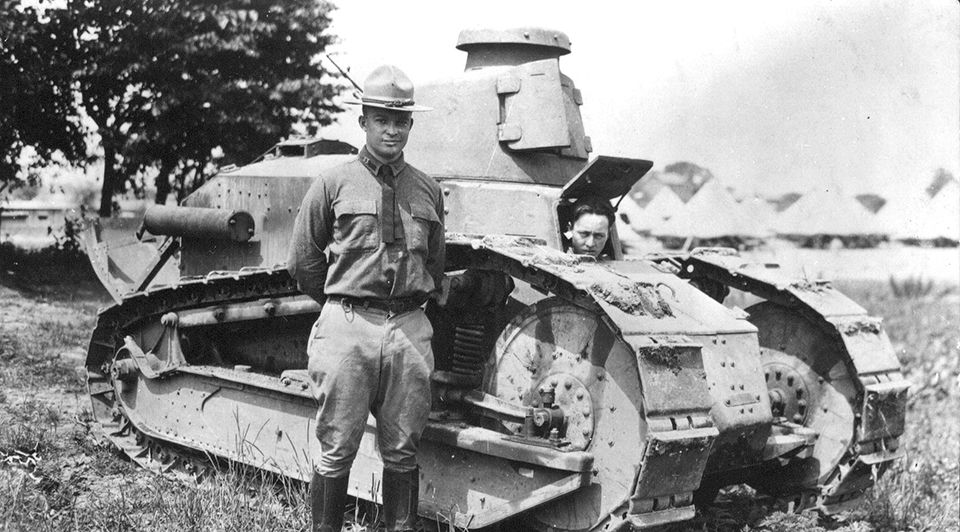 A black and white photograph of a young Dwight D. Eisenhower in a World War One uniform standing in front of a tank with a few trees and tents in the background.