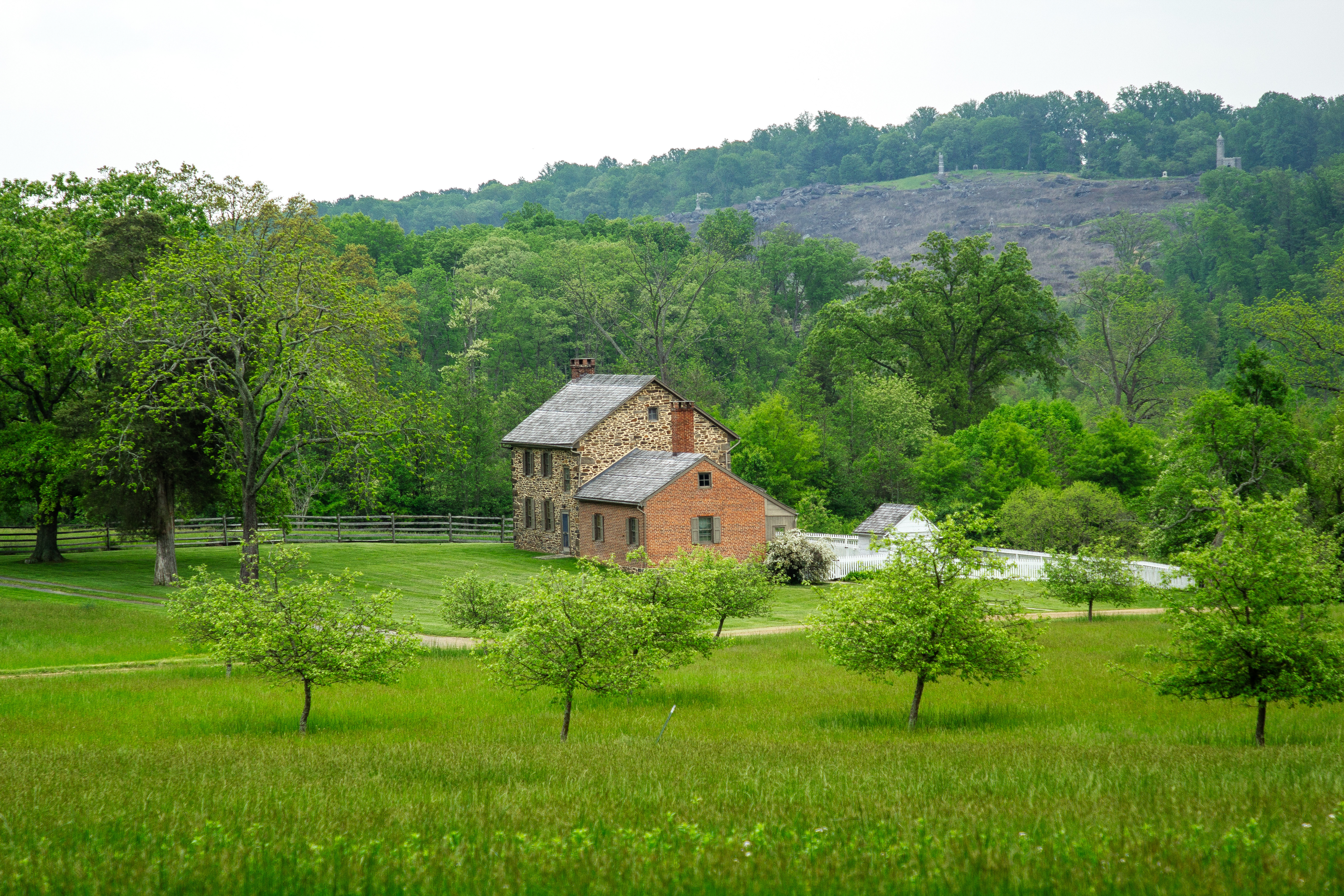 A stone and red brick house on grassy property with orchard trees sits in front of a heavily wooded area and a hill is in the distance.