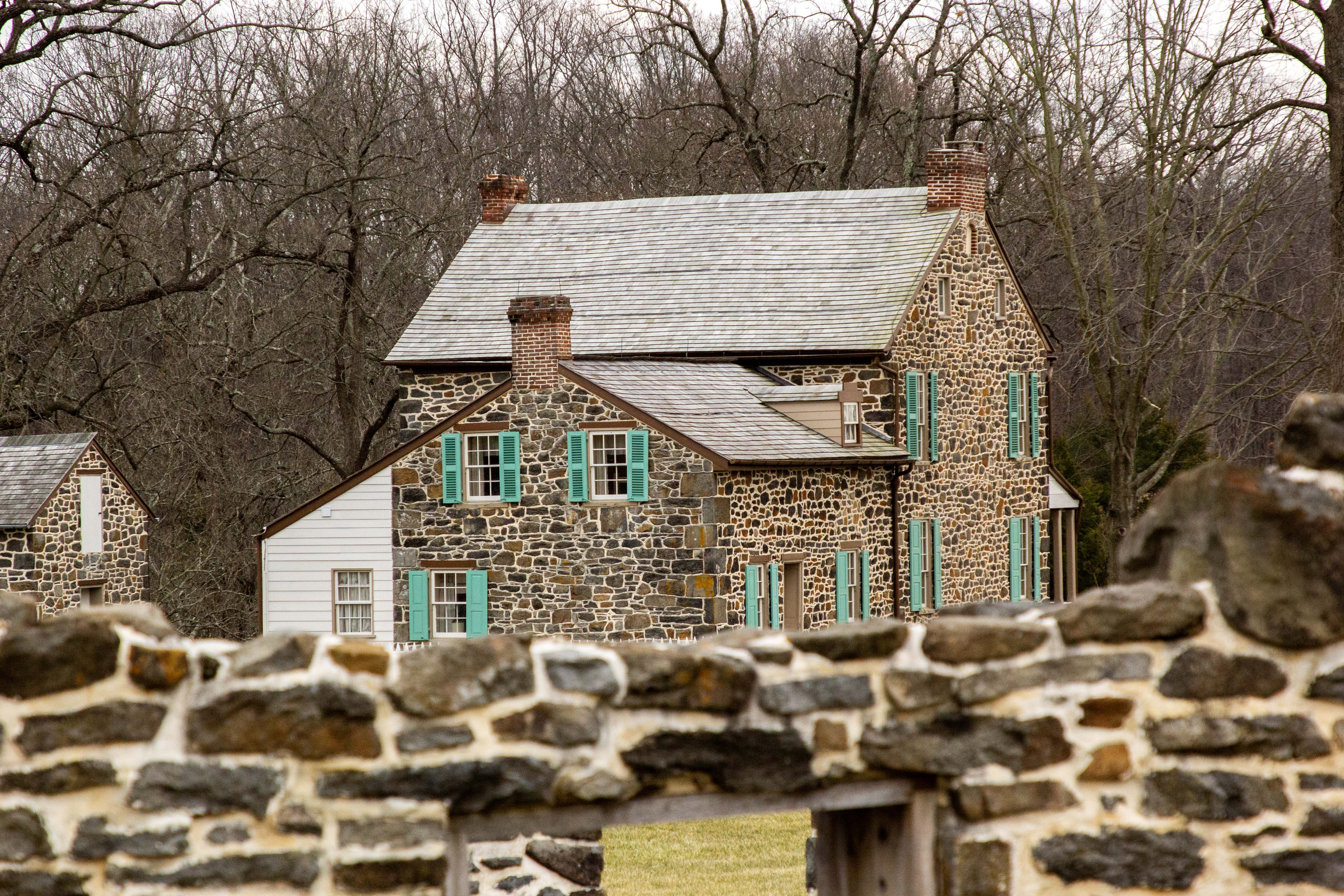 A two-story stone house with green shutters stands in front of a grove of trees with no foliage. The ruins of an old barn stand in the foreground.