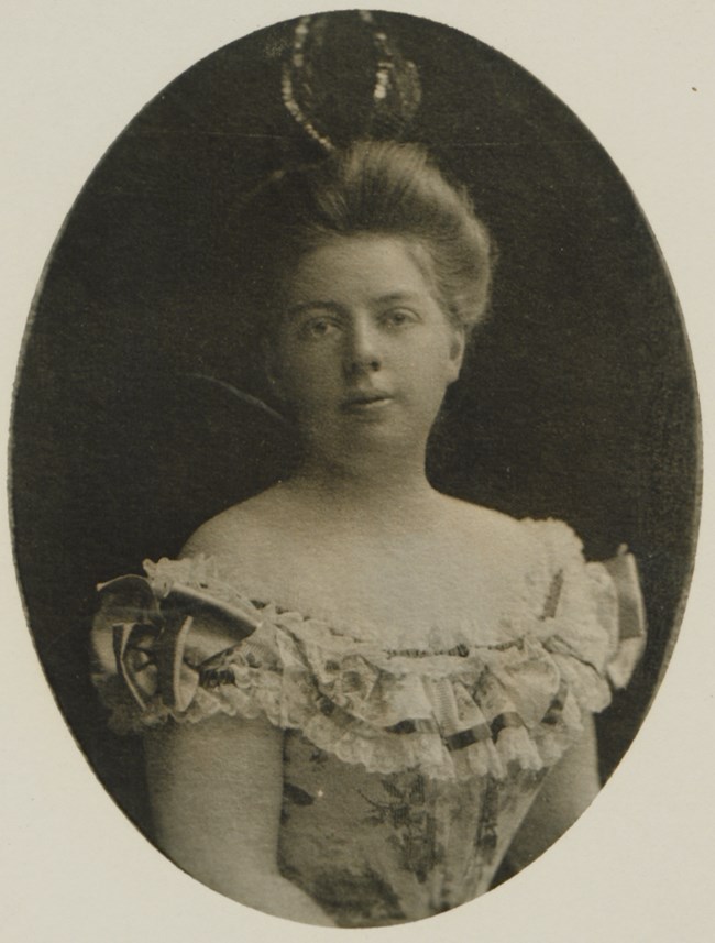 A black and white photo of a woman in a strapless dress from the 1890s with her hair pulled back with an elaborate headpiece on top.