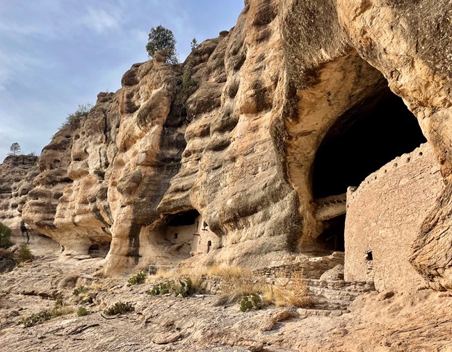 A view of the Gila Cliff Dwellings from outside the caves. The ancient stone structures are nestled within large natural caves, high on a cliffside. The surrounding landscape features rugged, rocky terrain with sparse vegetation