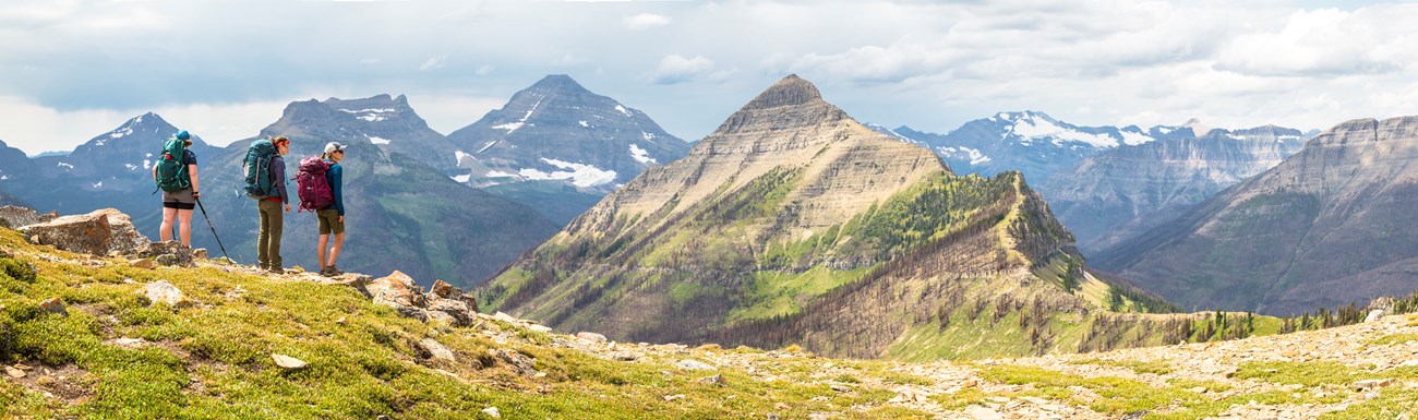 Three hikers stand in the foreground of a clearing on a mountain edge. Mountain peaks are seen in the distance with foliage or some snow still atop the ranges.