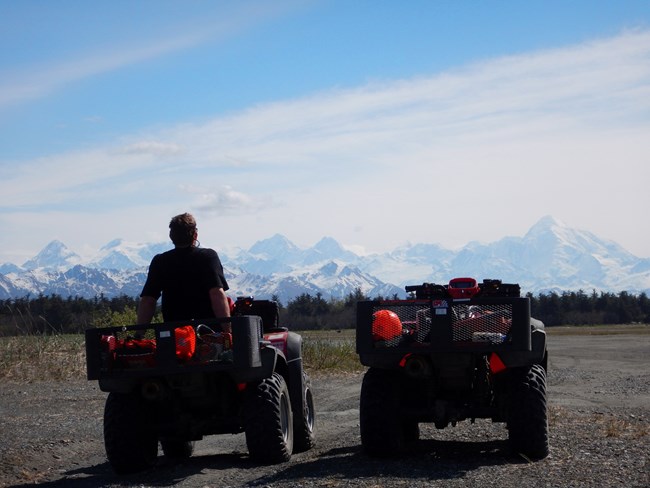 Two ATVs in front of snowy mountain range, one ATV has a person sitting on it.