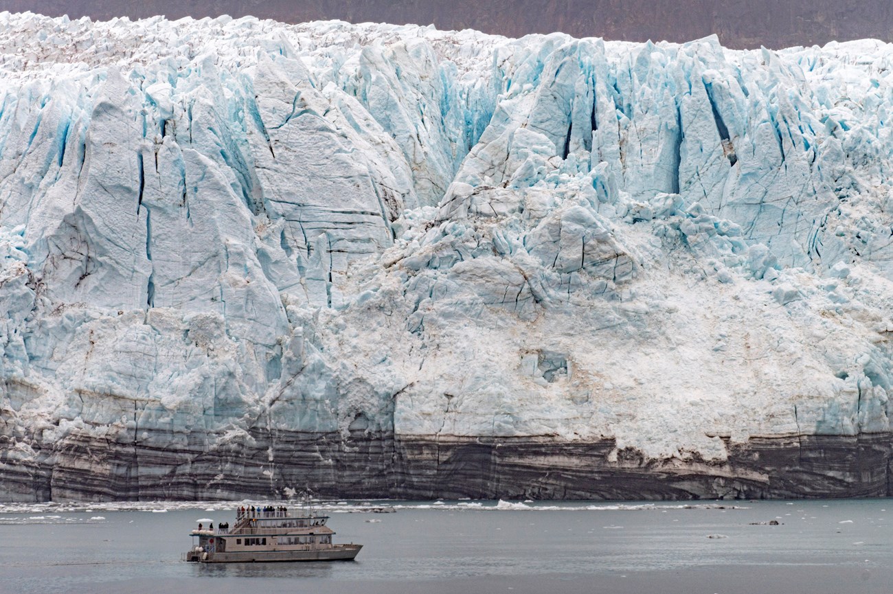 Day tour boat visit Margerie Glacier. A crumbly blue ice wall rises behind a metal catamaran-style boat.