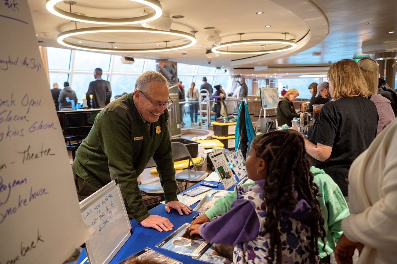A park ranger leans over a table to talk to a child. The table is filled with information cards about wildlife.