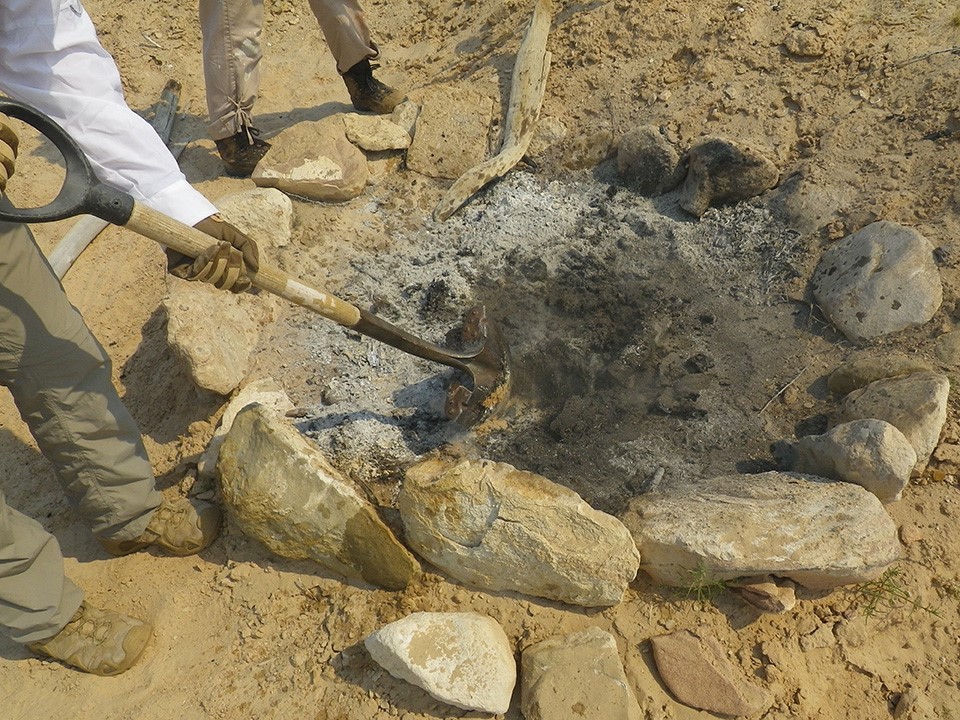 Ashes in fire ring on sandy area next to legs of two people. A shovel pokes at the ashes.