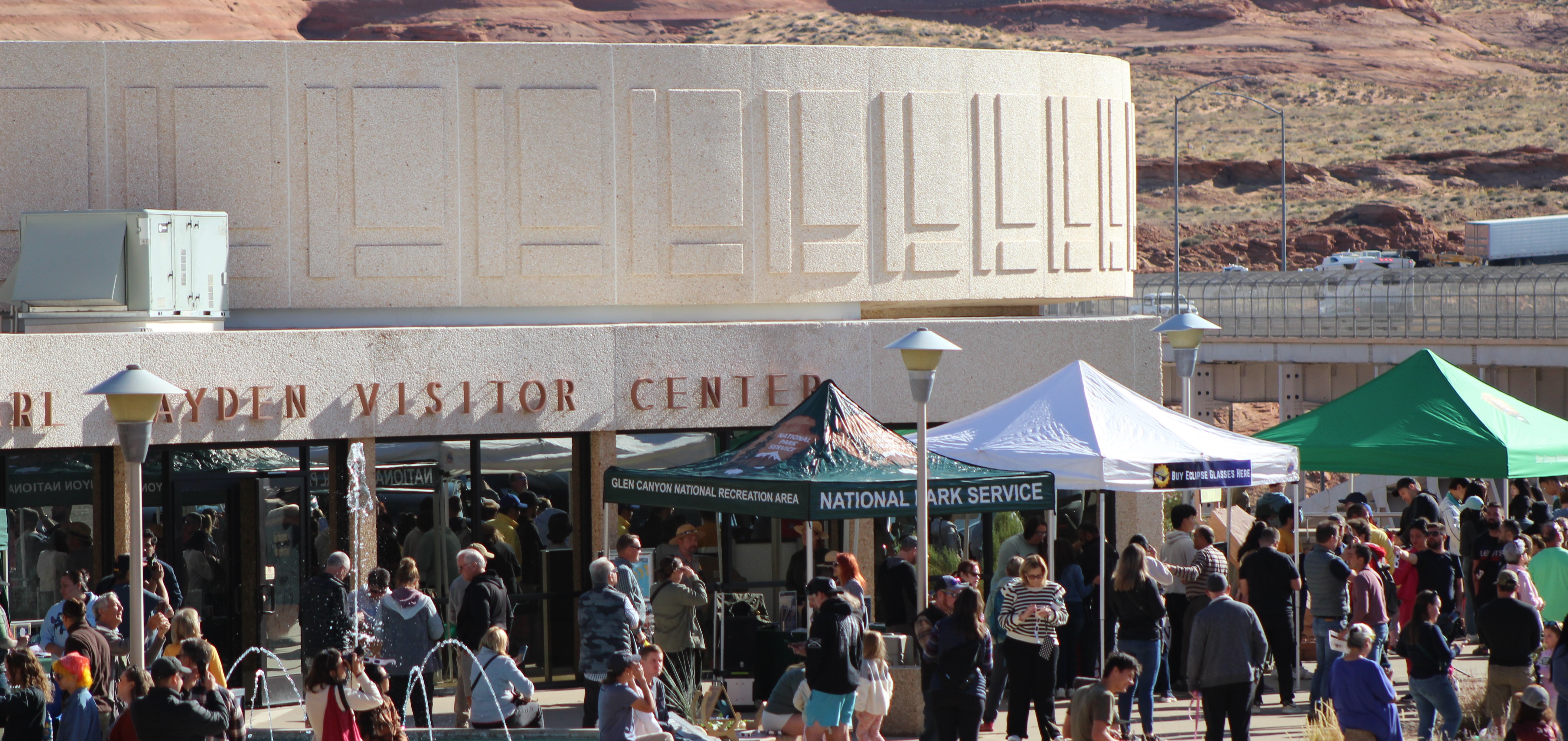 Crowd of people milling around a sidewalk in front of a concrete building with a circular edge.