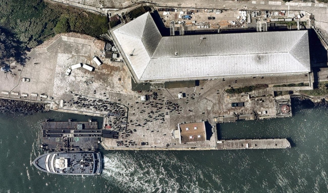 Color photo aerial view of Alcatraz concrete wharf with white ferry docked at lower right of picture. Large grey roof of building along top of photo. Dark green water of San Francisco Bay visible at bottom of photo.