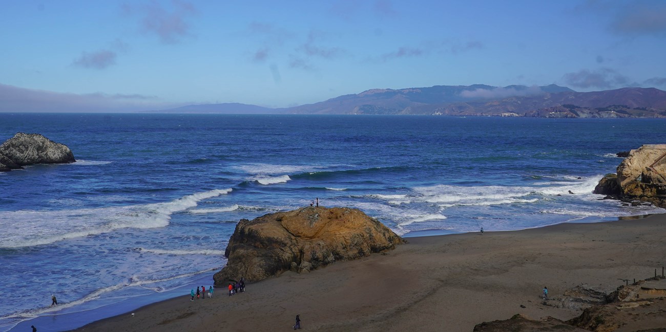 People walk, climb, and fish on a beach with large rocks and cliffs as waves crash on the shore.