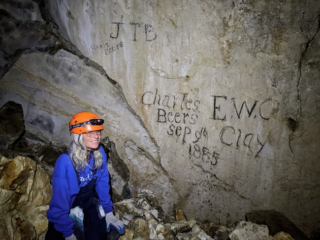 woman in blue cave suit and orange helmet sitting on rocks in front of flat rock wall with charcoal writing