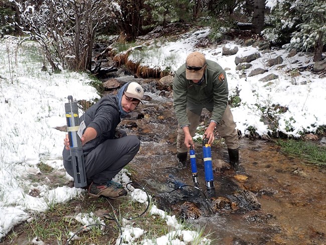 Two scientists sample water quality in a stream that has snow along its banks.