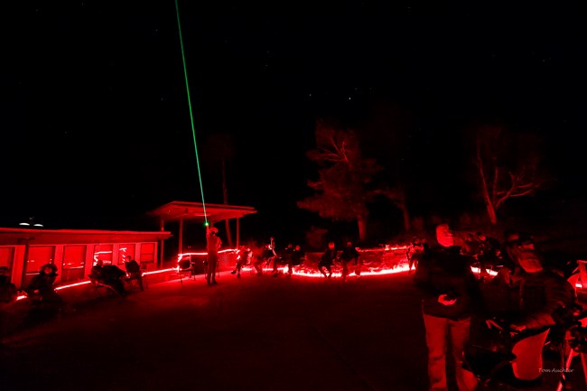 Surrounded by people and red lights, a park ranger points a bright green laser towards the sky. The sky itself is not pictured, but the small crowd seated in chairs surrounding the ranger is the focus.