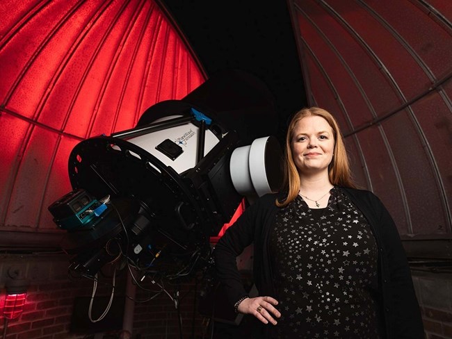 A blonde woman stands next to a black and white telescope under a red dome. She wears a black blouse.