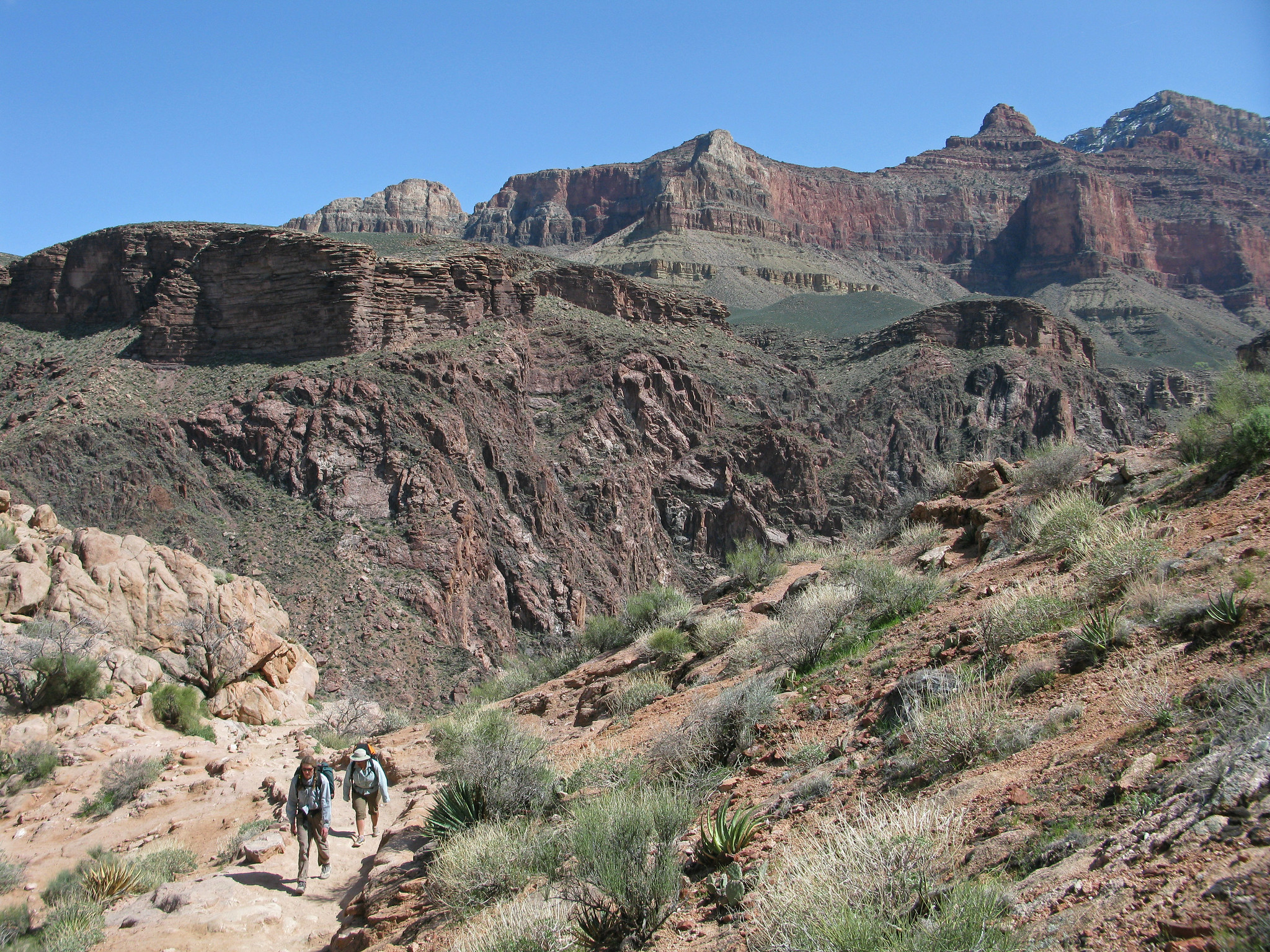 Two hikers with backpacks hike up the Bright Angel Trail with steep canyon walls in the background.