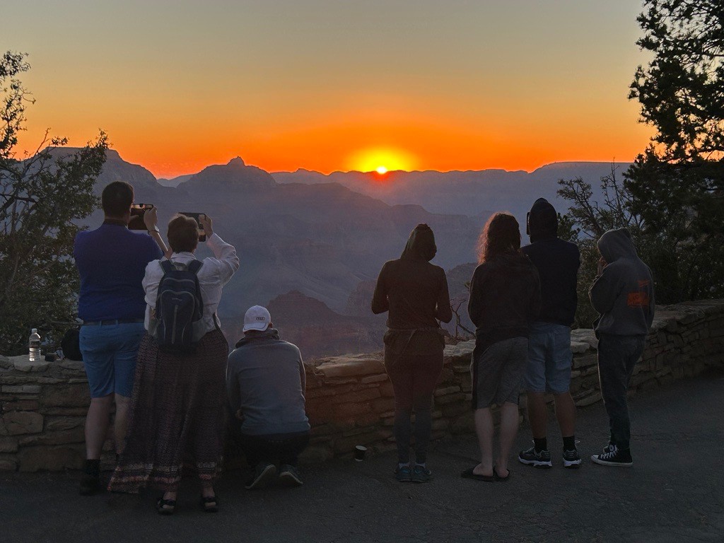 A group of visitors stands at the rim of the canyon taking photos of the sunrise