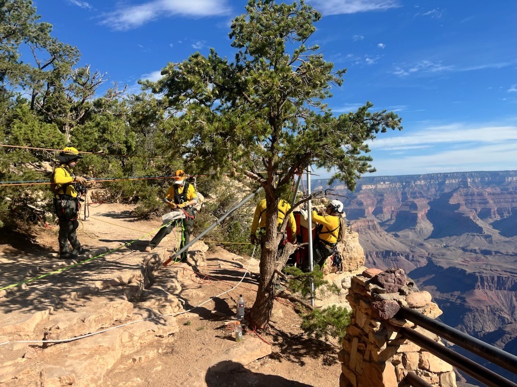 A team of six responders wearing yellow uniforms manage a technical rescue system at the edge of the canyon