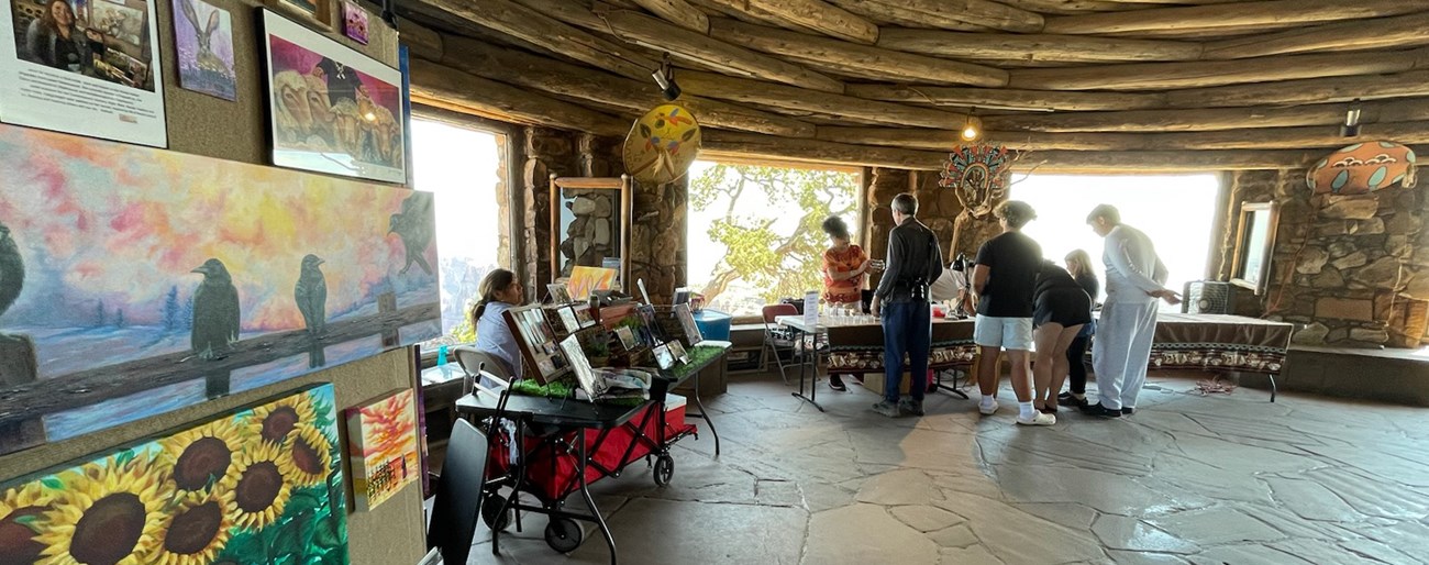 Colorful paintings of sheep, ravens, and sunflowers are placed on a display wall to the left. Next to it sits a few tables full of art with their artists sitting behind them talking with a group of visitors inside a curved room with a log ceiling.