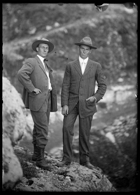 Historic black and white of two men standing wearing period suits and hats with trail switchbacks in the background