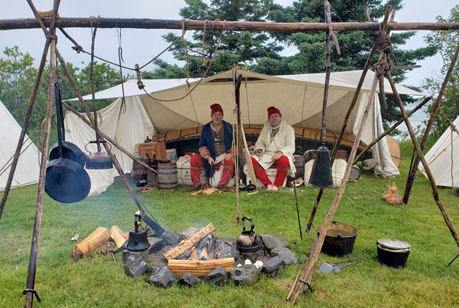 Two people in historic clothing, seated under a canvas tent with a historic cooking fire in the foreground.