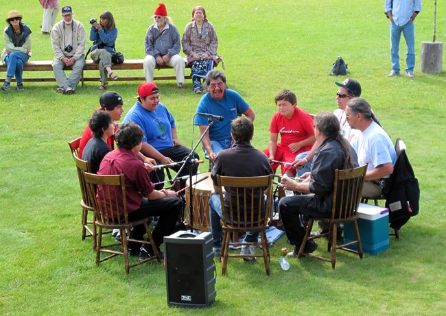 People seated in a circle around a drum with onlookers.