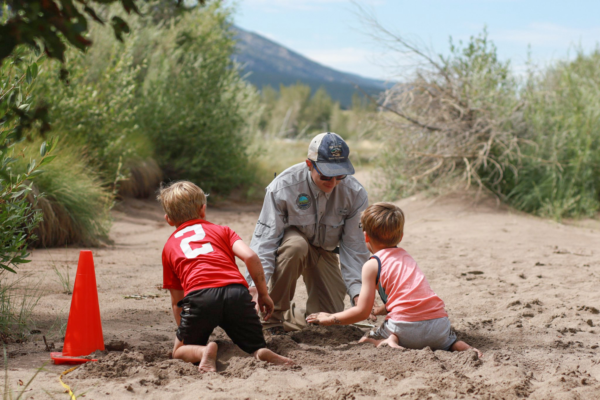 An intern in uniform helps two children in the sand during Junior Ranger Day 2022