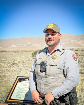 A man in a green ball cap and a grey park ranger uniform shirt standing in front of a view of the Sand Dunes