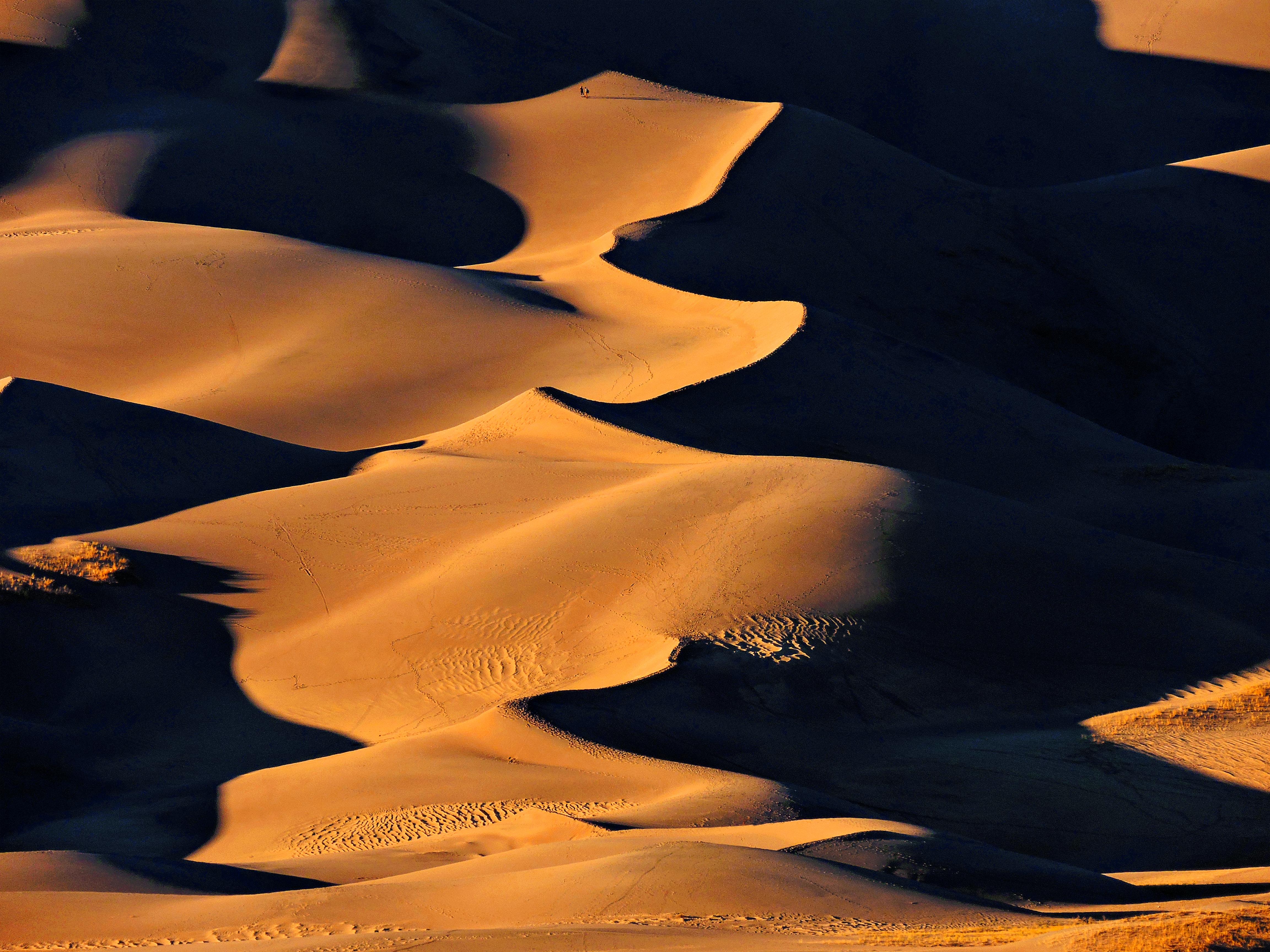 Two people walk on the Great Sand Dunes as shadows cover half the sand.