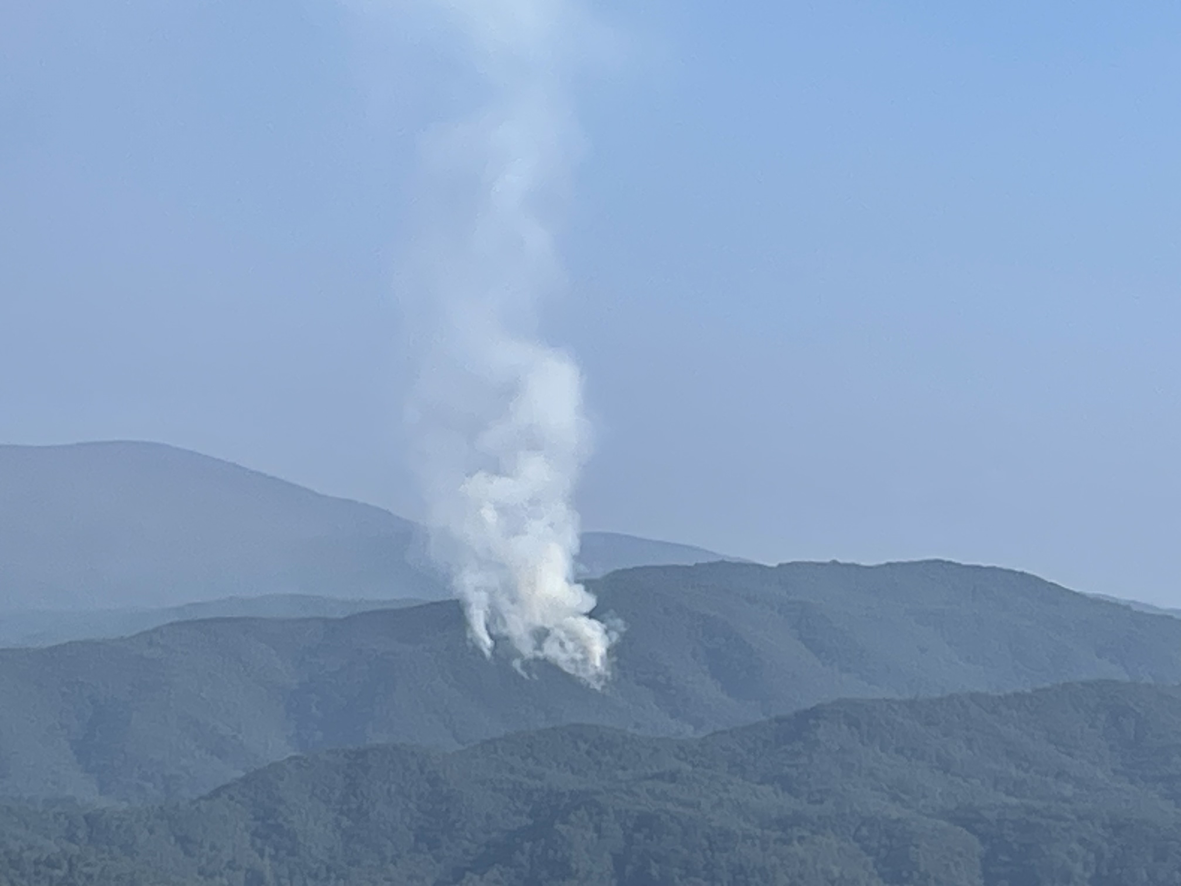 A plume of smoke rising from an area in rolling mountains beneath blue sky.