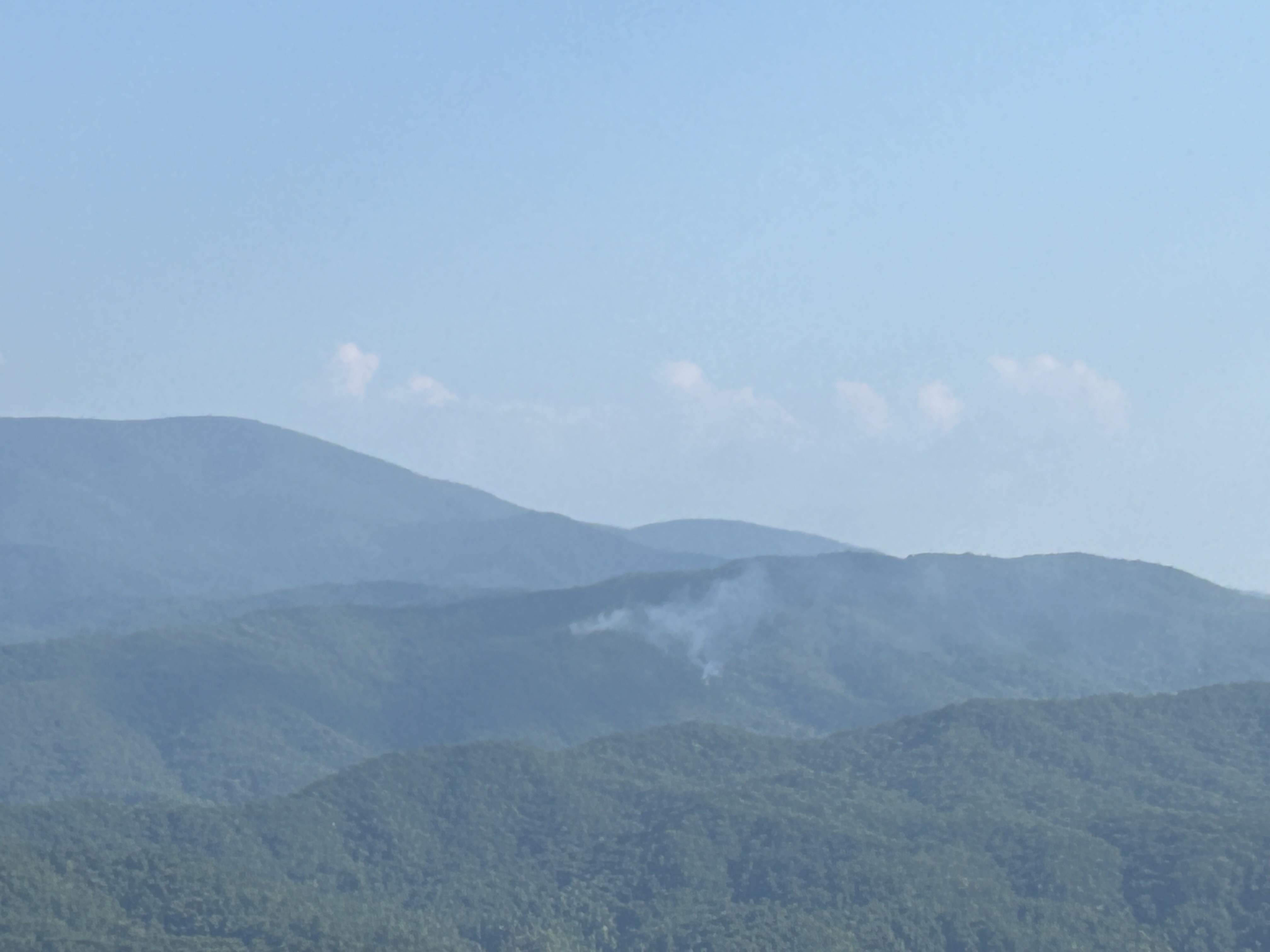 A small plume of smoke rising from between rolling mountain ridges on a mostly clear day.