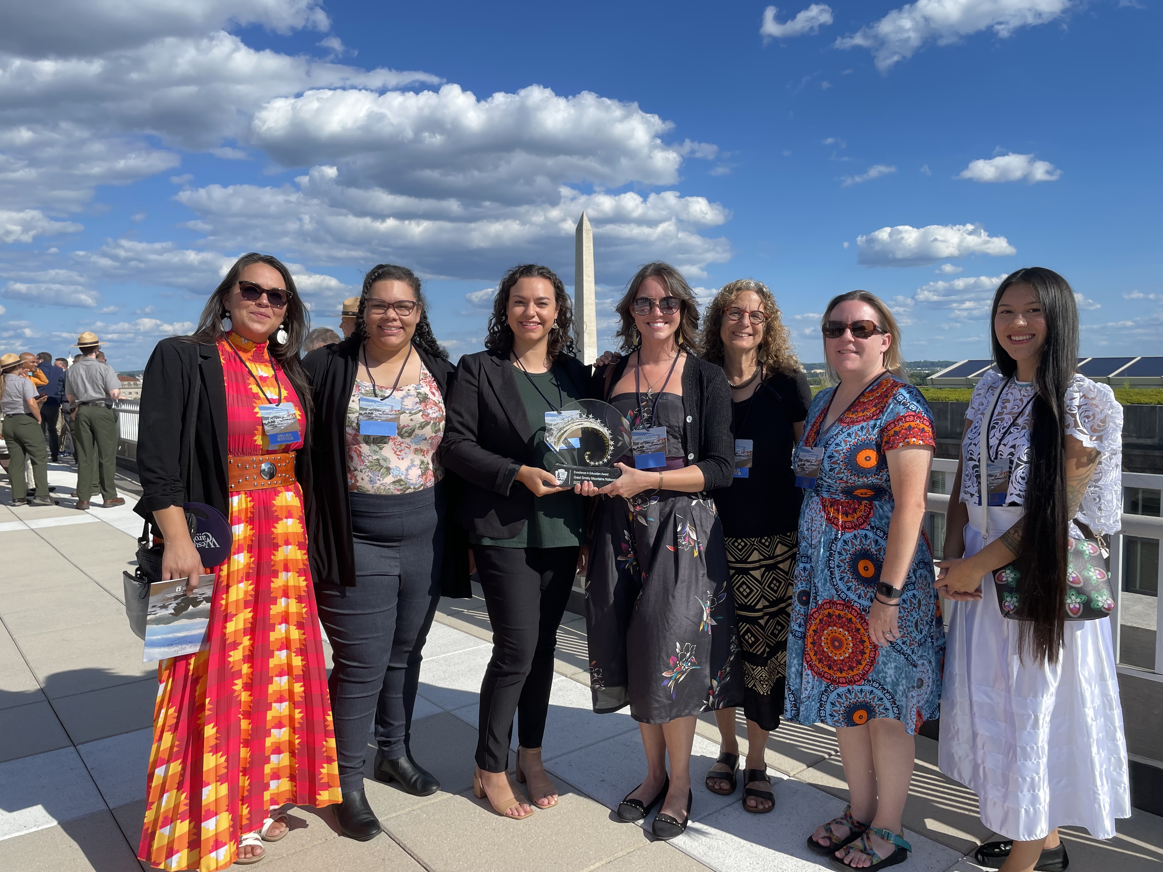 Seven people smiling in a row with the Washington Memorial in the background. The two center people hold a circular award. Park rangers and other people are in the left frame background.