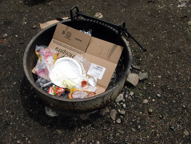 A firepit filled with wrappers, used plates and cutlery, carboard, and other abandoned trash.