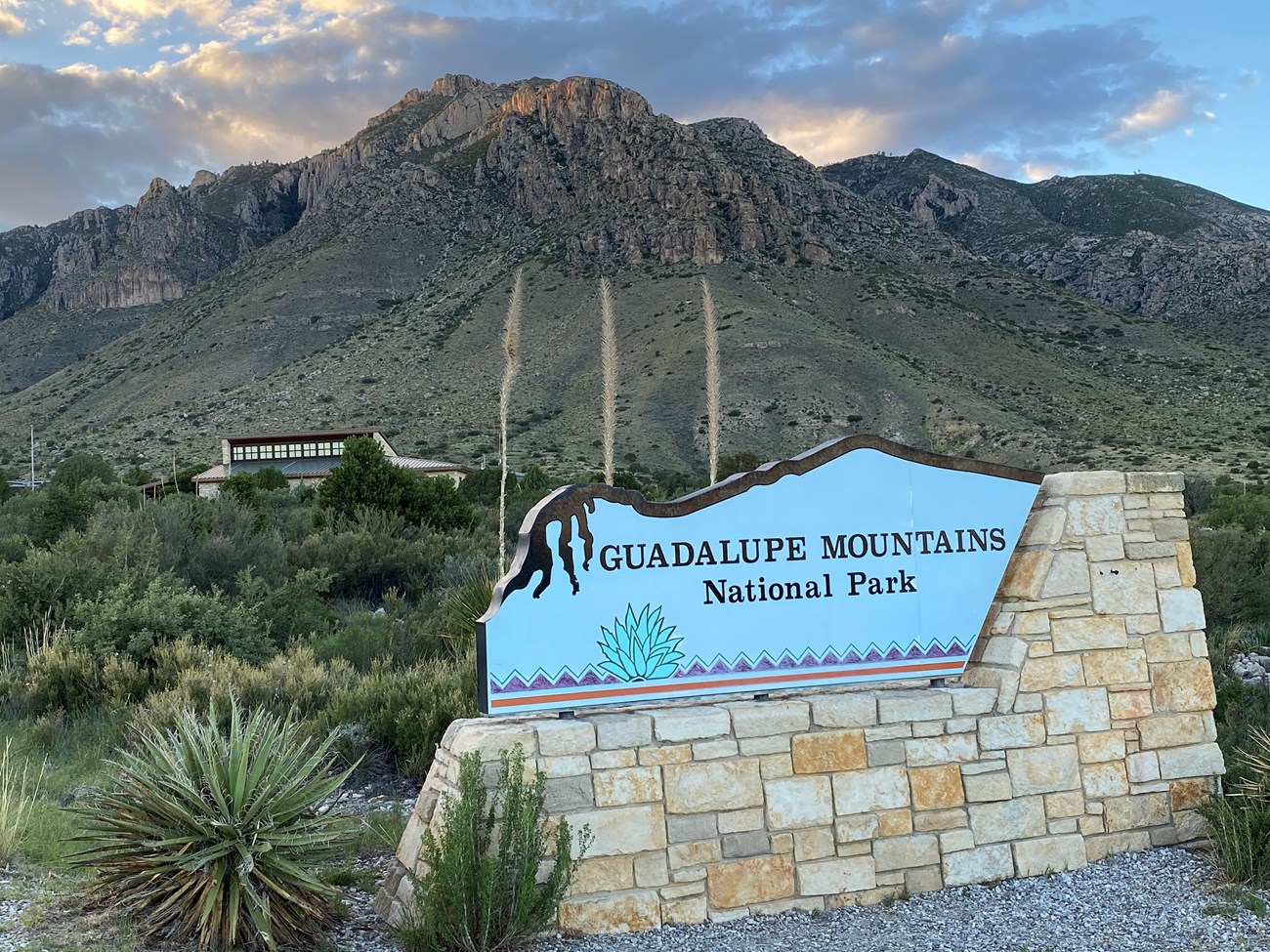 A park entrance sign stands before a building with high desert mounttains rising in the background.