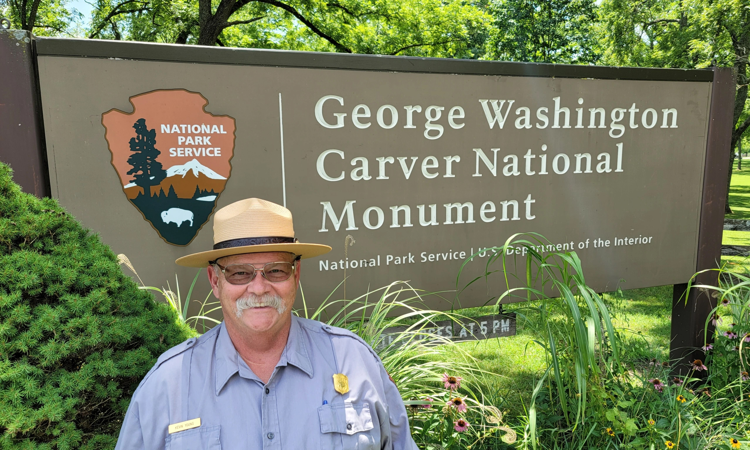 A smiling man in uniform with a white mustache stands in front of a sign for George Washington Carver National Monument.