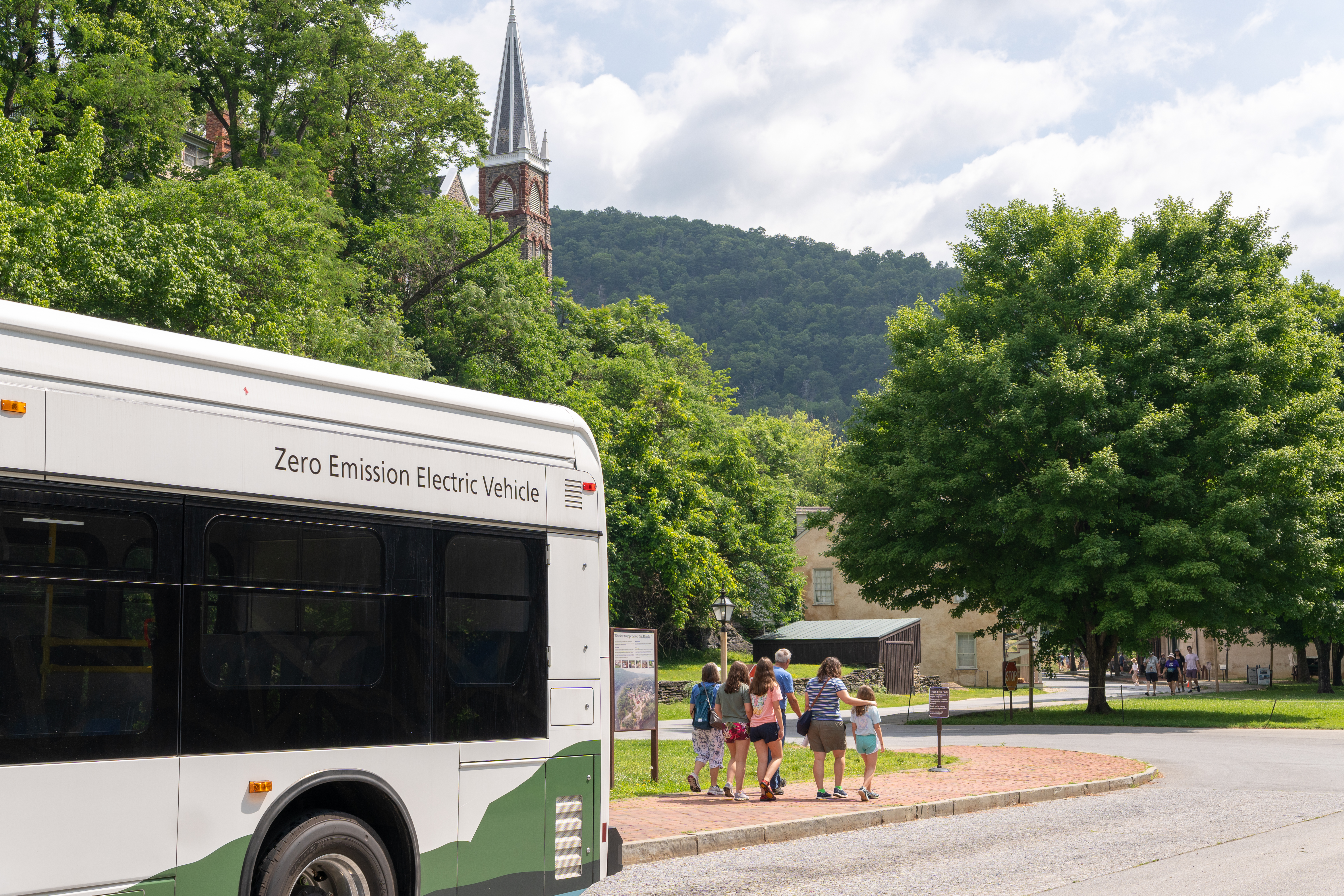 A family walks down a sidewalk towards a historic town after disembarking a shuttle bus..