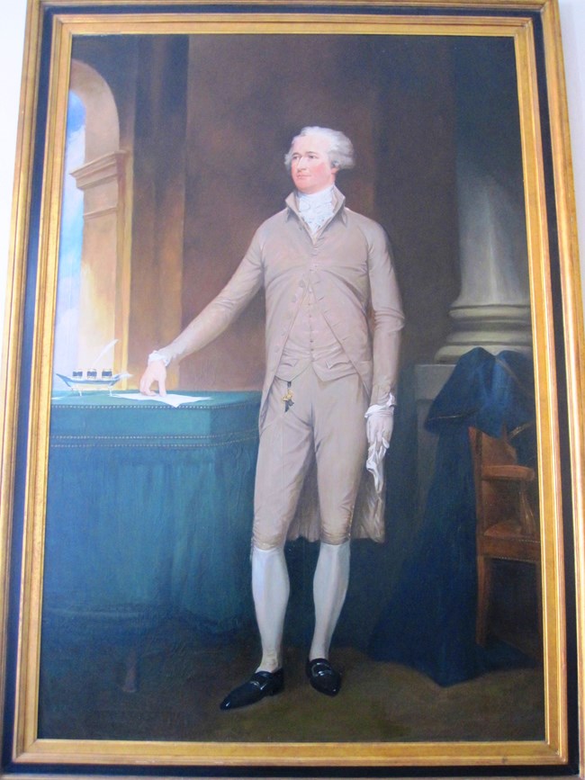 A full-length portrait of Alexander Hamilton, wearing a tan suit and resting one hand on a table next to an inkwell and parchment paper.